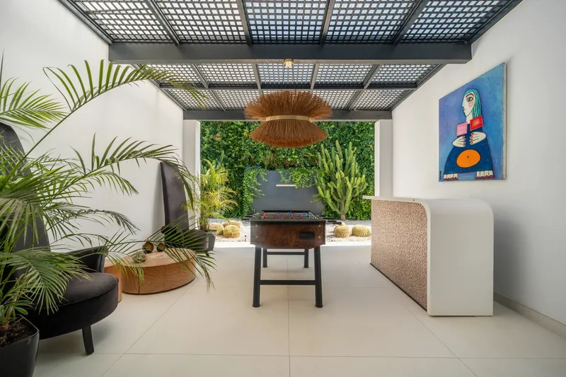Espace de jeux ombragé sous une pergola à lames, avec comptoir de bar et jardin de cactus