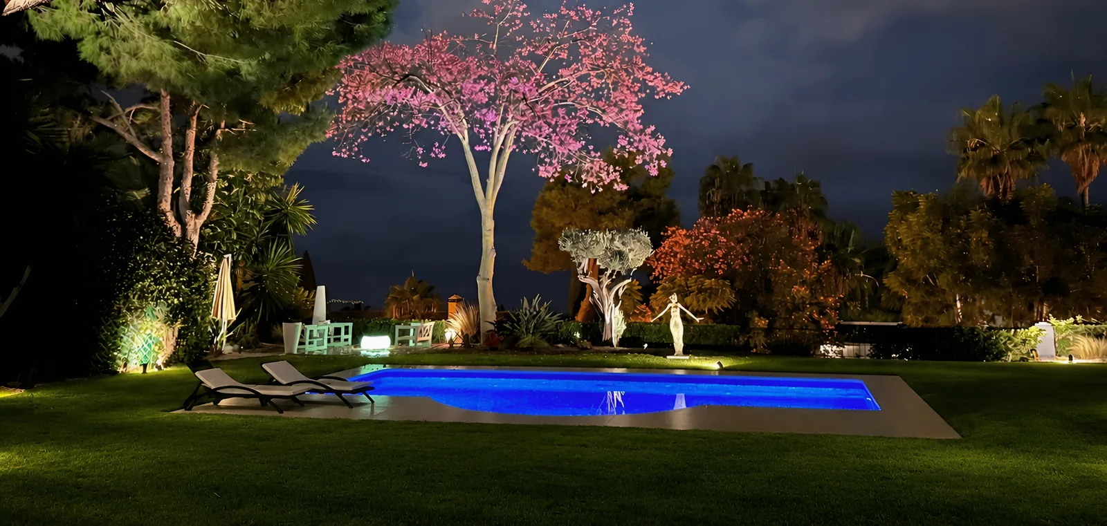 La piscine éclairée en bleu la nuit, avec des transats sur la pelouse et un arbre à fleurs roses illuminé au-dessus