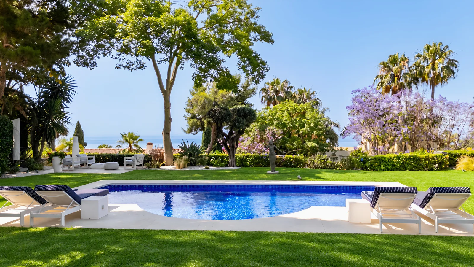 Transats au bord de la piscine, avec la pelouse, un jacaranda en fleurs et la Méditerranée au-delà du jardin