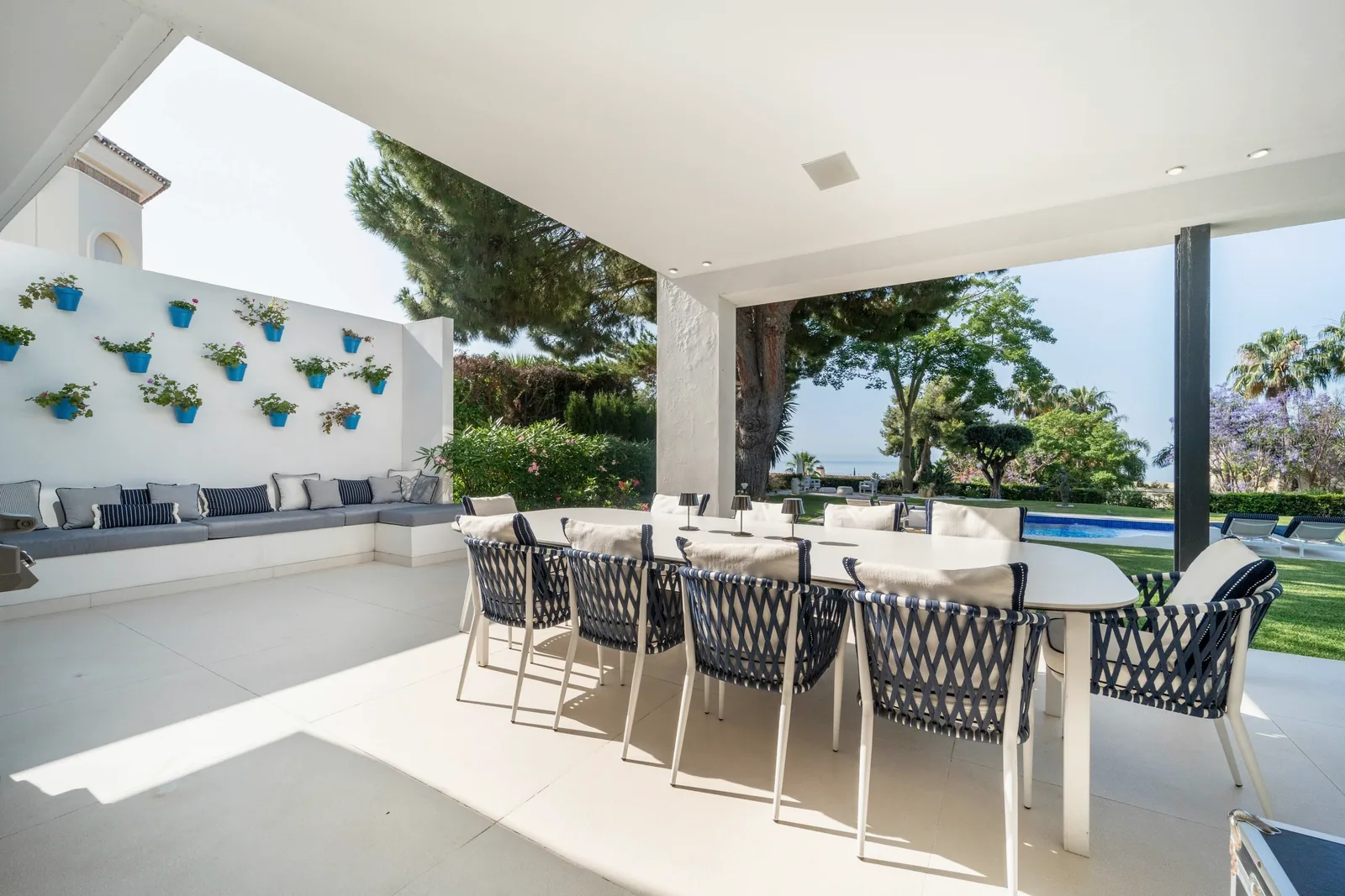 Terrasse couverte avec une table pour dix et des chaises en corde bleu marine, un mur de pots bleus, et la pelouse, la piscine et la mer au-delà